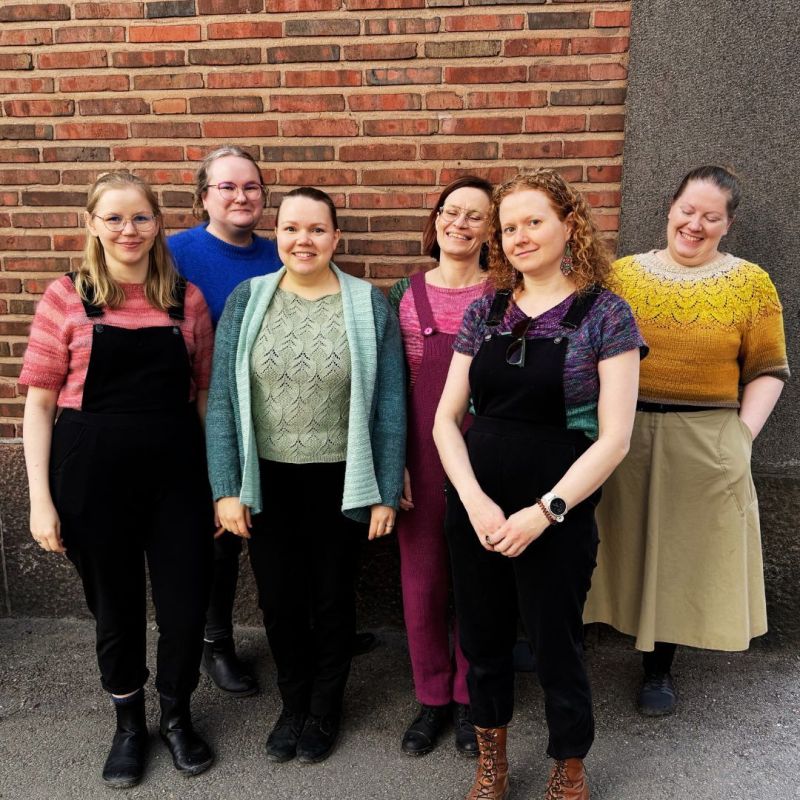 Kuvassa seisoo kuusi henkilöä ryhmänä tiiliseinän edustalla.